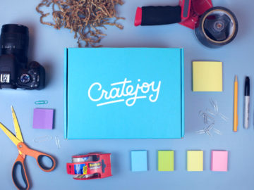 A closed Cratejoy box surrounded by a camera, a tape gun, various colors of Post-it notes, scissors, and a Sharpie marker.