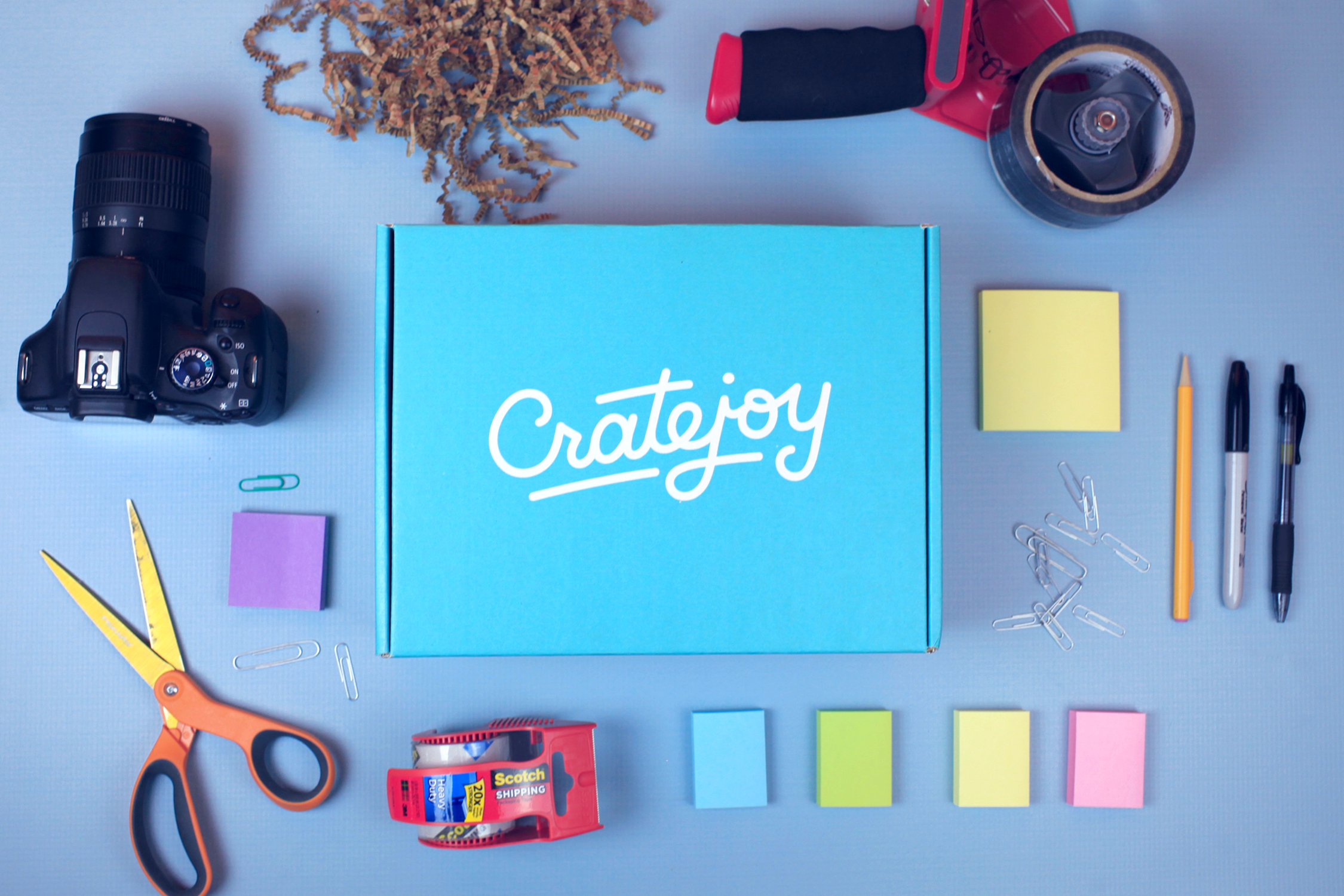 A closed Cratejoy box surrounded by a camera, a tape gun, various colors of Post-it notes, scissors, and a Sharpie marker.