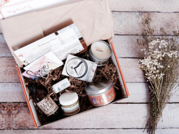 An open subscription box filled with a candle in a tin, a bottle of bath milk, encouraging cards and other self care products.