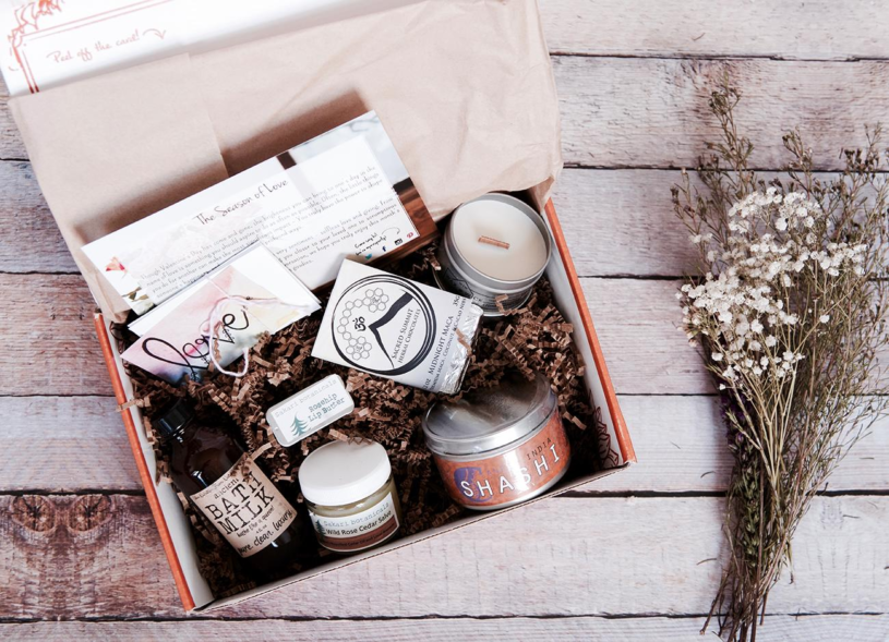 An open subscription box filled with a candle in a tin, a bottle of bath milk, encouraging cards and other self care products.
