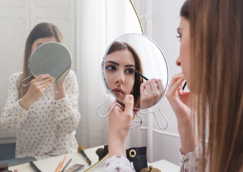 A woman sitting in front of a large mirror, holding a small mirror, putting on eye makeup.