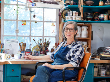 A woman sitting in a chair in a small art studio with painting supplies all around.